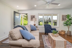 Ein Wohnzimmer mit einem beigefarbenen Sofa, einem dunklen Sessel, einem Deckenventilator und einem großen Fenster mit Blick auf eine grüne Landschaft.