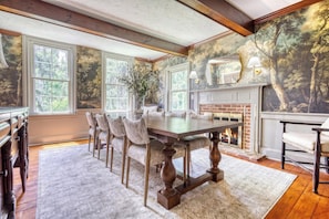 A dining room with a long table, chairs, a fireplace, and large windows.
