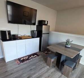 Cocina compacta con televisor de pantalla plana, microondas, cafetera y refrigerador. Cuenta con una pequeña mesa de comedor con un banco y una planta en maceta.