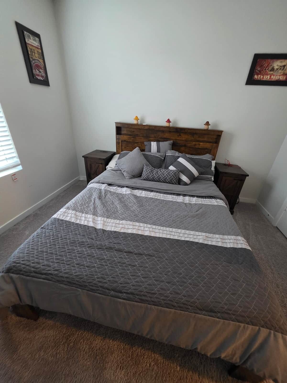 A bedroom with a wooden headboard, a bed with grey bedding, two nightstands, and a window with blinds.
