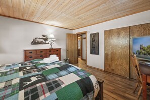 Bedroom view showing a wood-plank ceiling, plaid deer-print quilt, large wooden dresser, sliding closet doors, desk with TV, and en-suite doorway. - Big Bear Hot Tub | Game Room + Putting Green (Big Bear Lake)