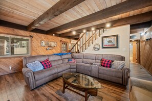 Large brown sectional sofa with throw pillows, oval glass coffee table, wood-paneled walls, exposed ceiling beams, and staircase. - Big Bear Hot Tub | Game Room + Putting Green (Big Bear Lake)