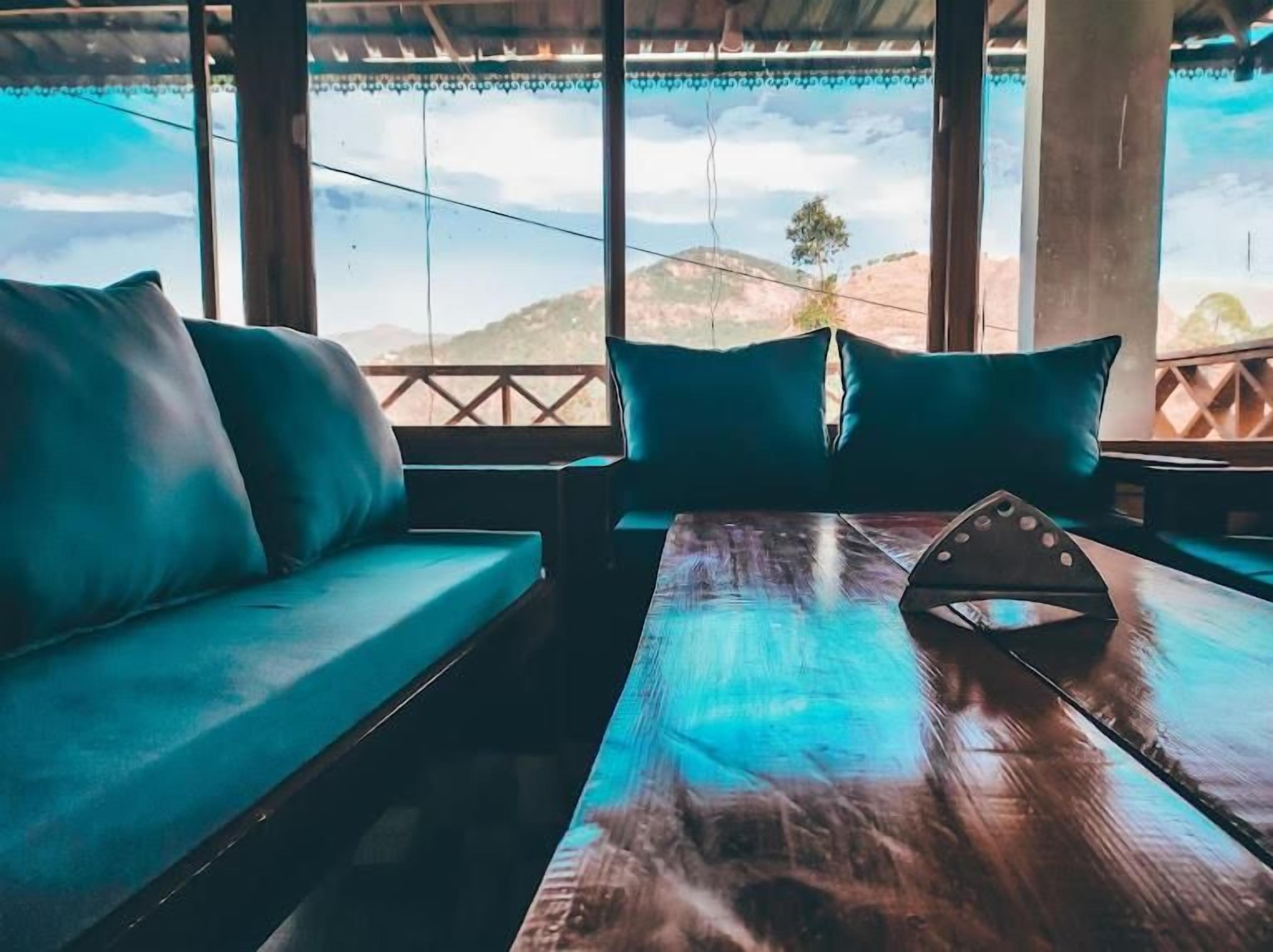 A wooden table with a triangular object in the center, a blue sofa with cushions, and a view of hills through large windows.