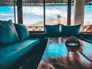A wooden table with a triangular object in the center, a blue cushioned bench with a pillow, and a view of hills through large windows. - Shiva Valley Resort (Dhanaulti)
