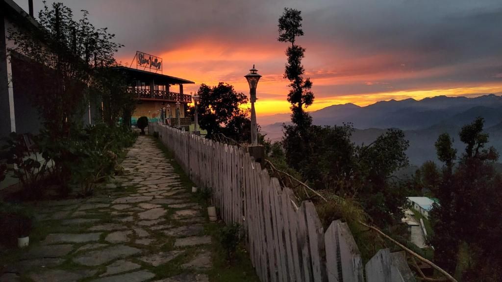 A stone pathway leading to a building with a wooden fence, surrounded by trees and mountains in the background.