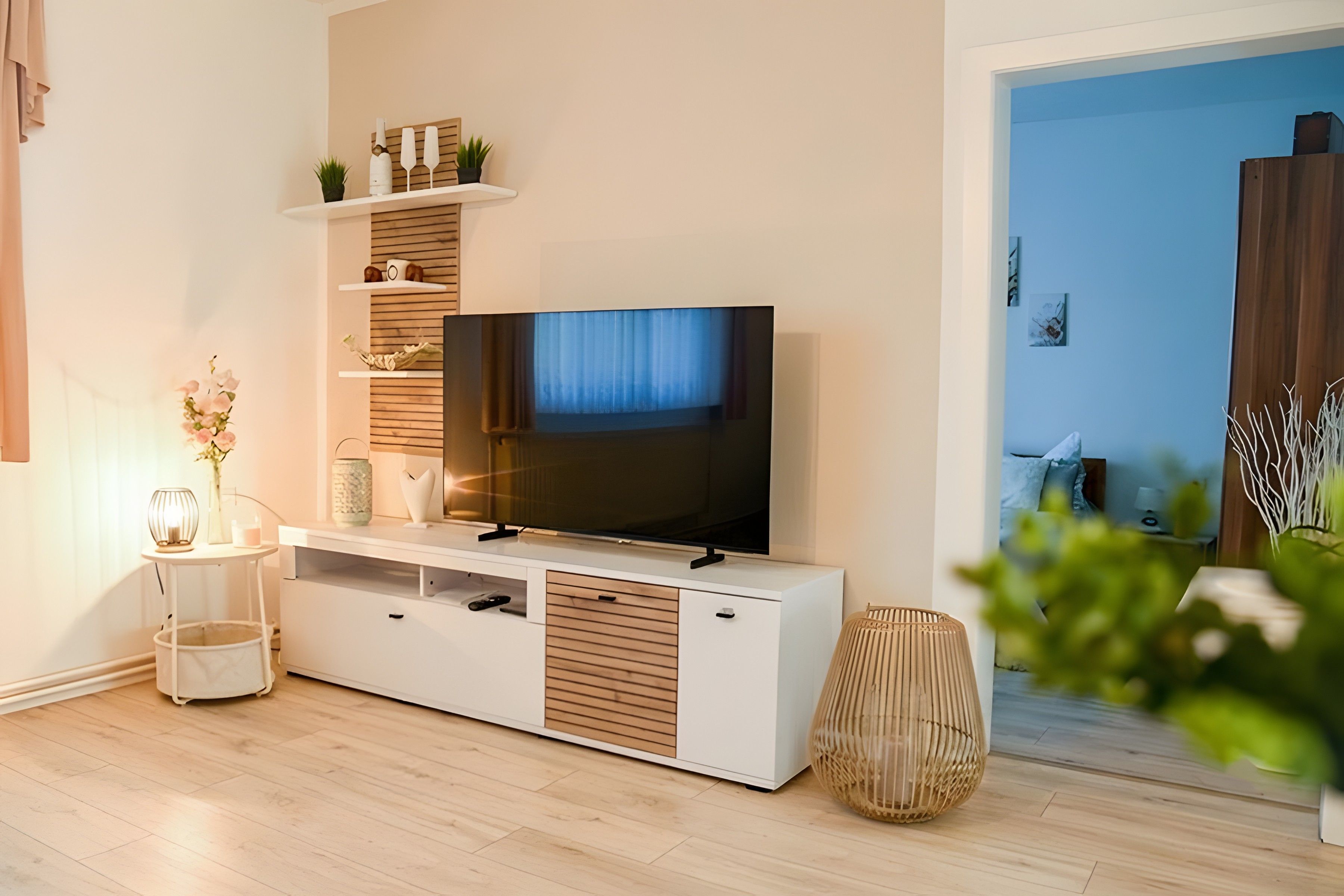 Ein modernes Wohnzimmer mit einem Flachbildfernseher auf einer weißen Unterhaltungsecke, einem kleinen Beistelltisch mit Lampe und einem Topfpflanzen.