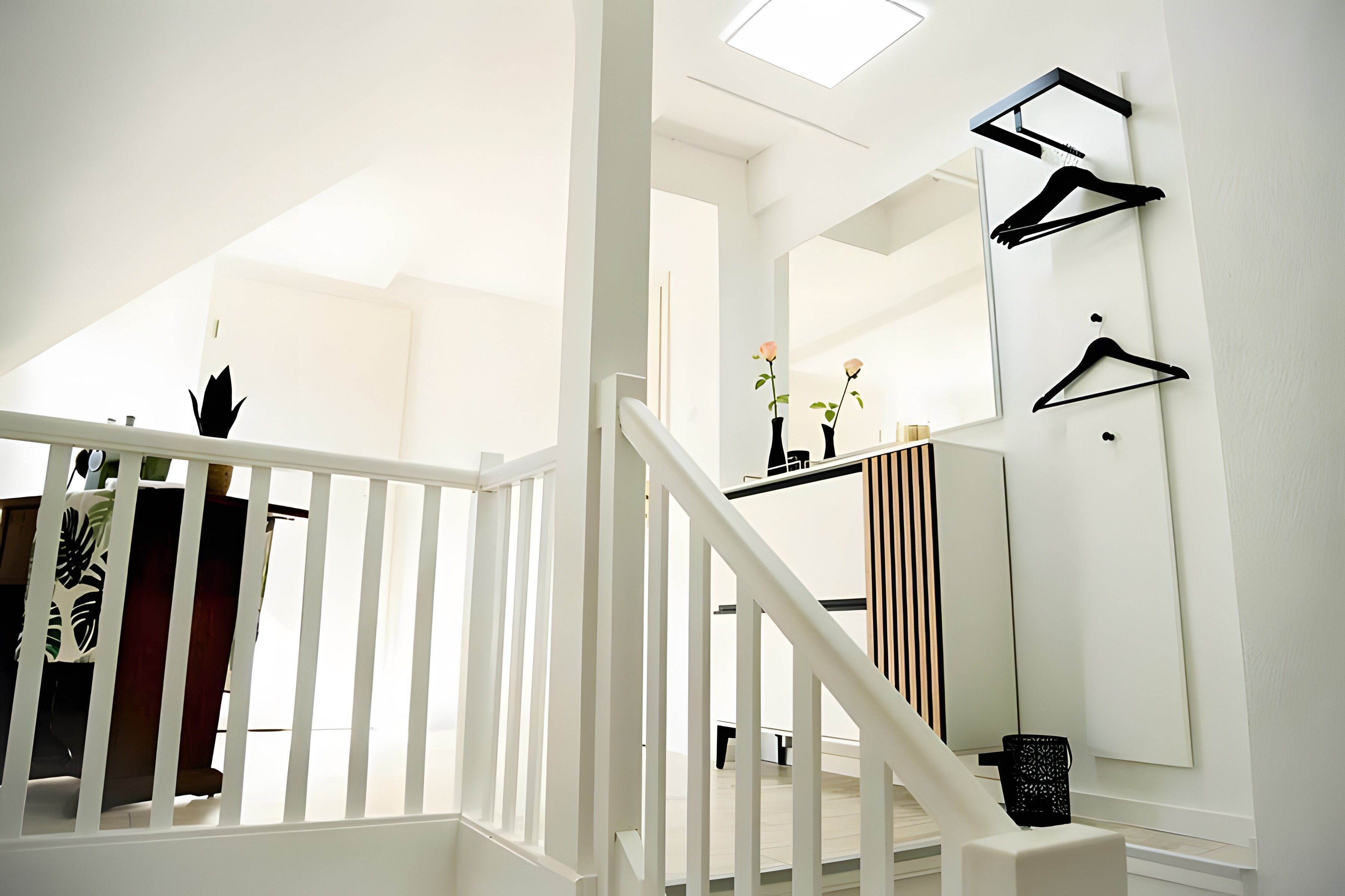 Un escalier avec des rampes blanches menant à un palier agrémenté d’un miroir et d’un porte-manteau noir.
