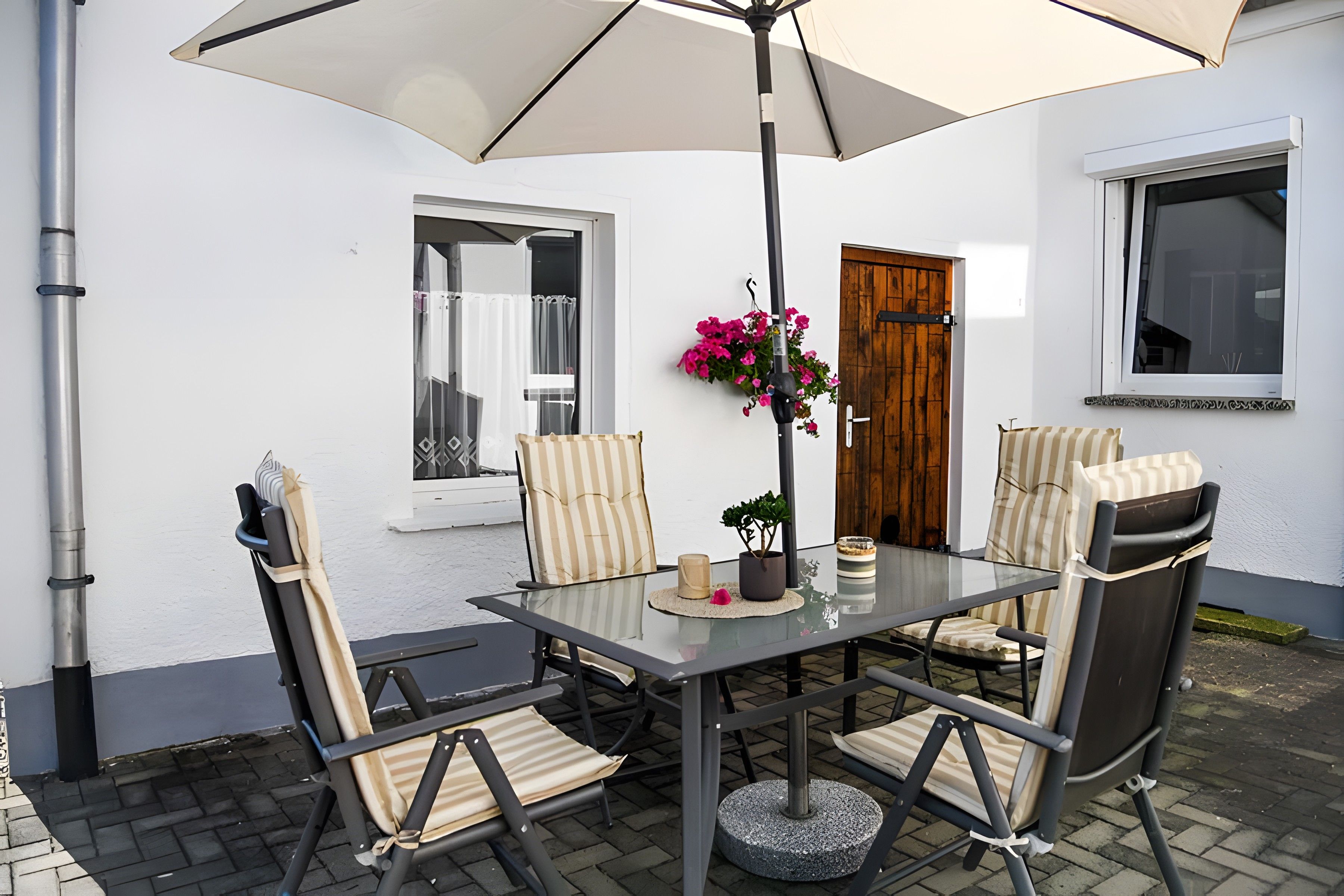 Eine Terrasse im Freien mit einem Glastisch, gestreiften Stühlen, einer hängenden Blumenarragemang und einer Holztür.