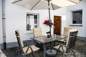 Un patio al aire libre con una mesa de vidrio, sillas a rayas, un arreglo floral colgante y una puerta de madera.