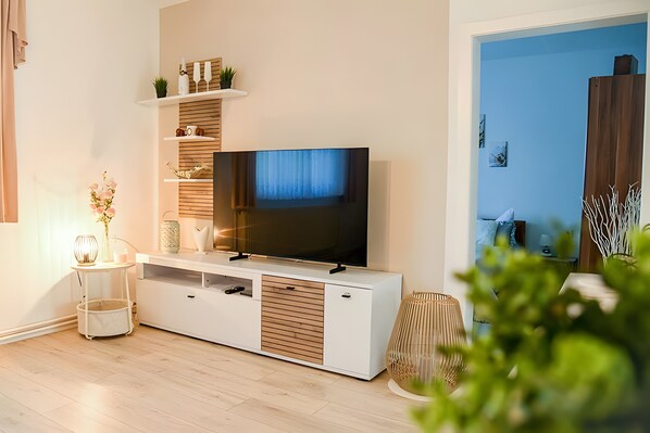 A modern living room with a flat-screen TV, wooden shelves, a small table with a vase, and a potted plant.