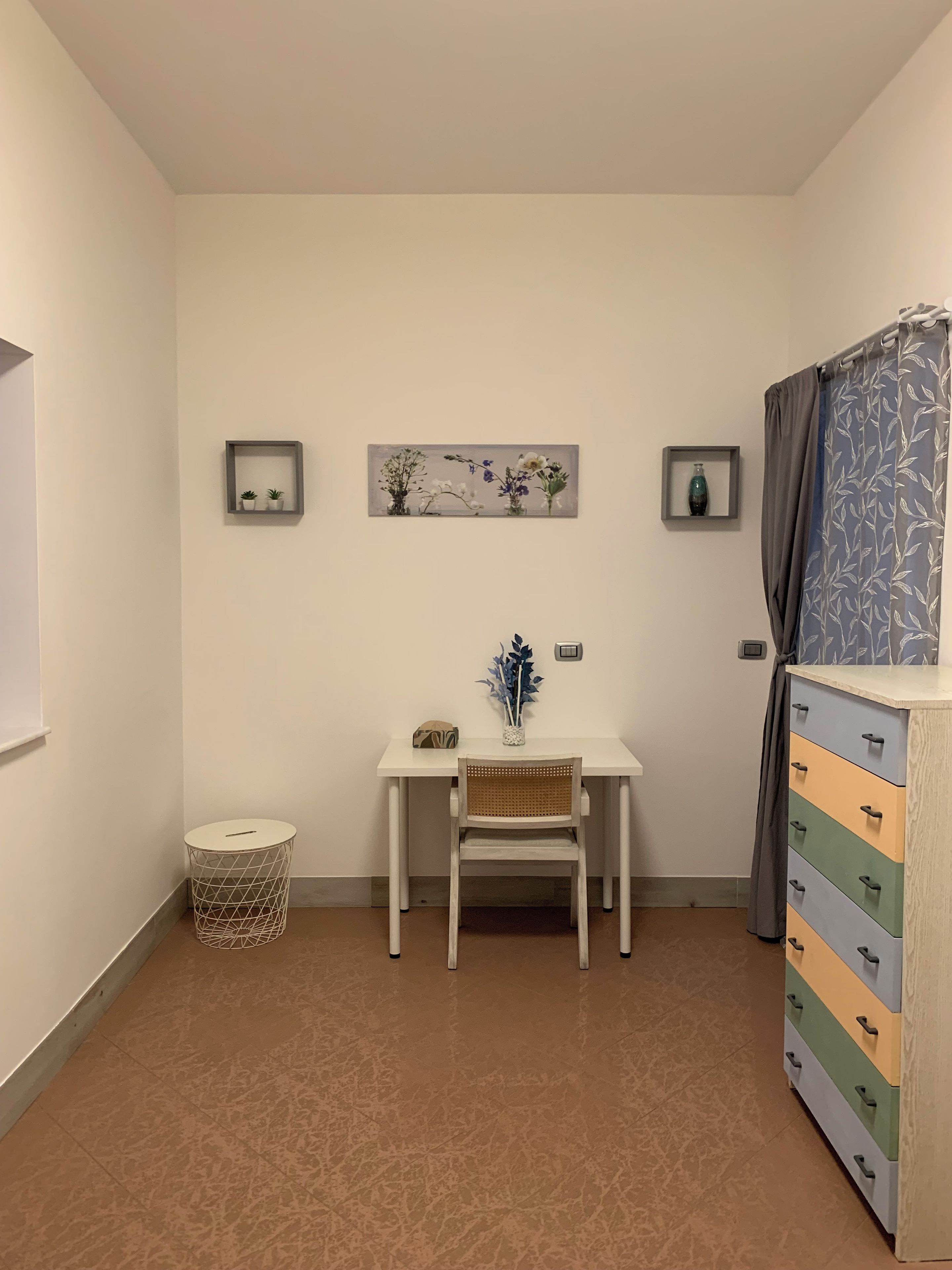 Een kamer met een wit bureau, een stoel, een klein rond tafeltje, een kast met lades en twee wandplanken met decoratieve objecten.