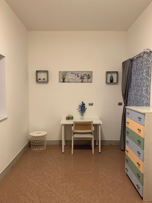 A room with a white desk, a chair, a small round table, a cabinet with drawers, and two wall-mounted shelves with decorative items.