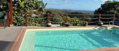 A swimming pool with a wooden deck, surrounded by a rope fence, and overlooking a landscape with hills and a view of the sea.