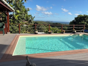 A swimming pool with a wooden deck, surrounded by a rope fence, and overlooking a landscape with hills and a view of the sea. - Ground-floor studio in a rural setting overlooking the sea (Le François)