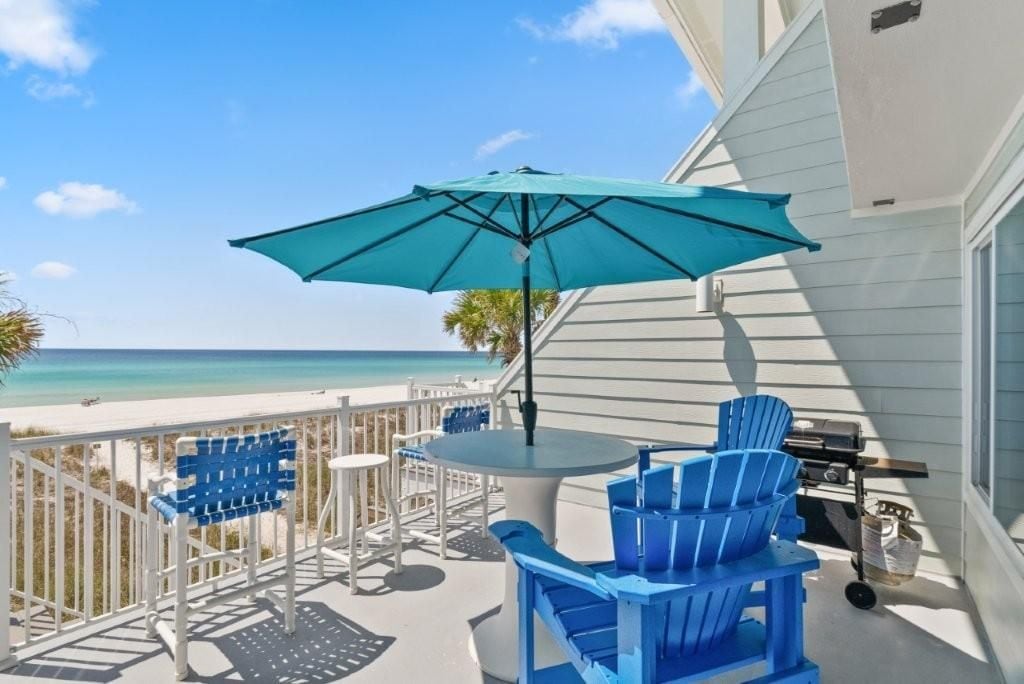 Un balcon avec vue sur la plage, des chaises bleues, une table et un barbecue.