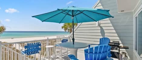A balcony with a view of the beach, blue chairs, a table, and a grill.