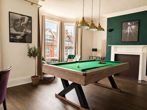 A pool table with a green felt surface, surrounded by wooden flooring, in a room with a fireplace, a green accent wall, and a window with curtains. - Cromer Coastal Escape | 2 Mins to Beach, Pool Table & Games Room | Sleeps 11 (Cromer)