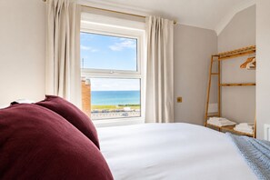 A bedroom with a bed, a window with curtains, a view of the sea, and a wooden shelf with towels. - Cromer Coastal Escape | 2 Mins to Beach, Pool Table & Games Room | Sleeps 11 (Cromer)