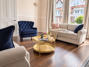 A living room with a blue armchair, a beige sofa, and a round gold coffee table with a book and a vase. - Cromer Coastal Escape | 2 Mins to Beach, Pool Table & Games Room | Sleeps 11 (Cromer)