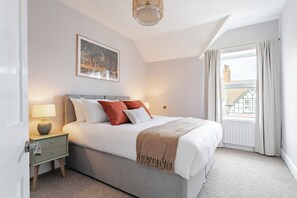 A hotel room with a bed, a nightstand, a lamp, a painting on the wall, and a window with curtains. - Cromer Coastal Escape | 2 Mins to Beach, Pool Table & Games Room | Sleeps 11 (Cromer)
