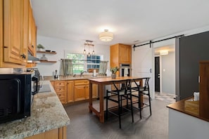 A modern kitchen with wooden cabinets, a central island with bar stools, a black microwave, and a hanging light fixture. - Lake Cumberland Farmhouse: Arcade Room, Fire Pit, Boat Parking, Pet Friendly (Russell Springs)
