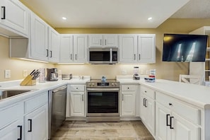 A modern kitchen with white cabinetry, a built-in microwave, an oven, and a flat-screen TV mounted on the wall. - 🎣 Palm Court Yacht Club 312A | Waterfront Views, Pool & Fishing Dock 🌊 (Fort Walton Beach)