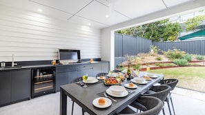 A modern outdoor kitchen with a dining table set for a meal, a grill, and a view of a garden. - Gloucester Escape, 37a Gloucester St - stunning duplex with fabulous water views (Nelson Bay)