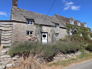 A stone cottage with a white door, multiple windows, and a stone wall. - Farthing Cottage (Wareham)