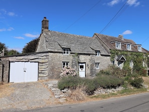 A stone house with a white garage door, a chimney, and a small garden with shrubs. - Farthing Cottage (Wareham)