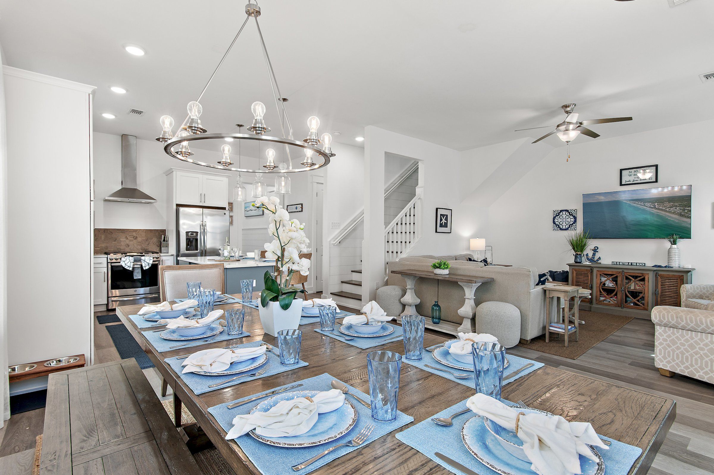 A modern kitchen with stainless steel appliances, a dining area set for six with blue placemats and white napkins, and a living room with a large painting and a ceiling fan.