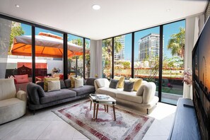A modern living room with a large window, a sofa set, a coffee table, and a TV. - Duplex 3BR with Garden Terrace & Glass Lounge (Dubai)