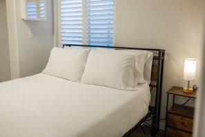Uma cama com roupa de cama branca, uma mesa de cabeceira com abajur e uma janela com persianas.