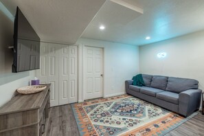 Quarto com sofá cinzento, mesa lateral de madeira, tapete estampado e TV de tela plana fixada na parede.