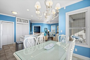 A modern kitchen with white cabinetry, a dining area with a glass table, and a living room with a sofa and a painting. - Oceanfront 2BR Condo | Pool + Covered Deck + Easy Beach Access (Pine Knoll Shores)