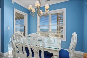A dining area with a white table and chairs, a chandelier, and large windows with blinds. - Oceanfront 2BR Condo | Pool + Covered Deck + Easy Beach Access (Pine Knoll Shores)