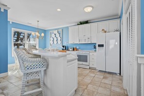 A kitchen with white cabinetry, a built-in island, and a dining area with wicker chairs. - Oceanfront 2BR Condo | Pool + Covered Deck + Easy Beach Access (Pine Knoll Shores)
