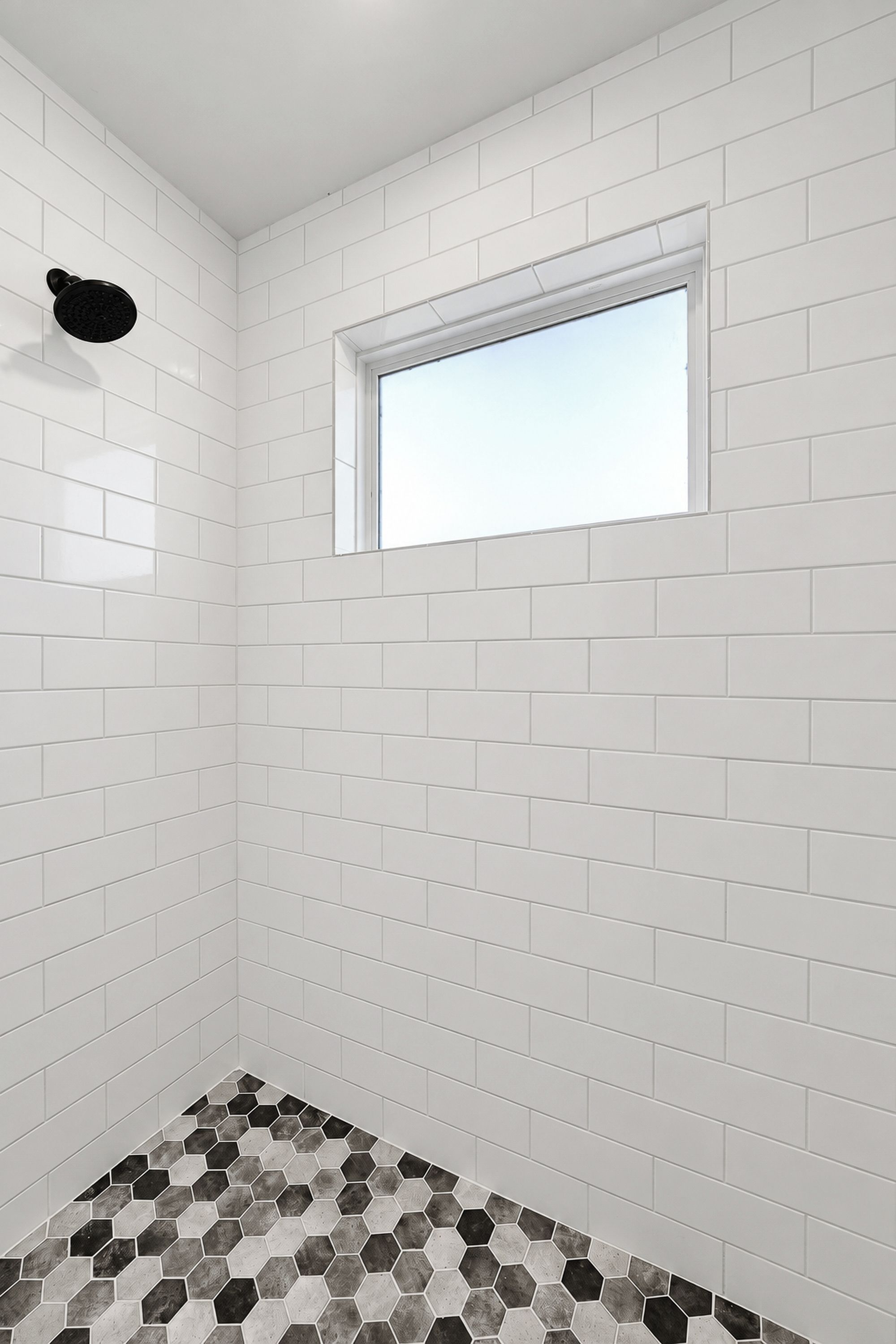 A bathroom with white subway tiles, a black showerhead, a window, and a hexagonal patterned floor.