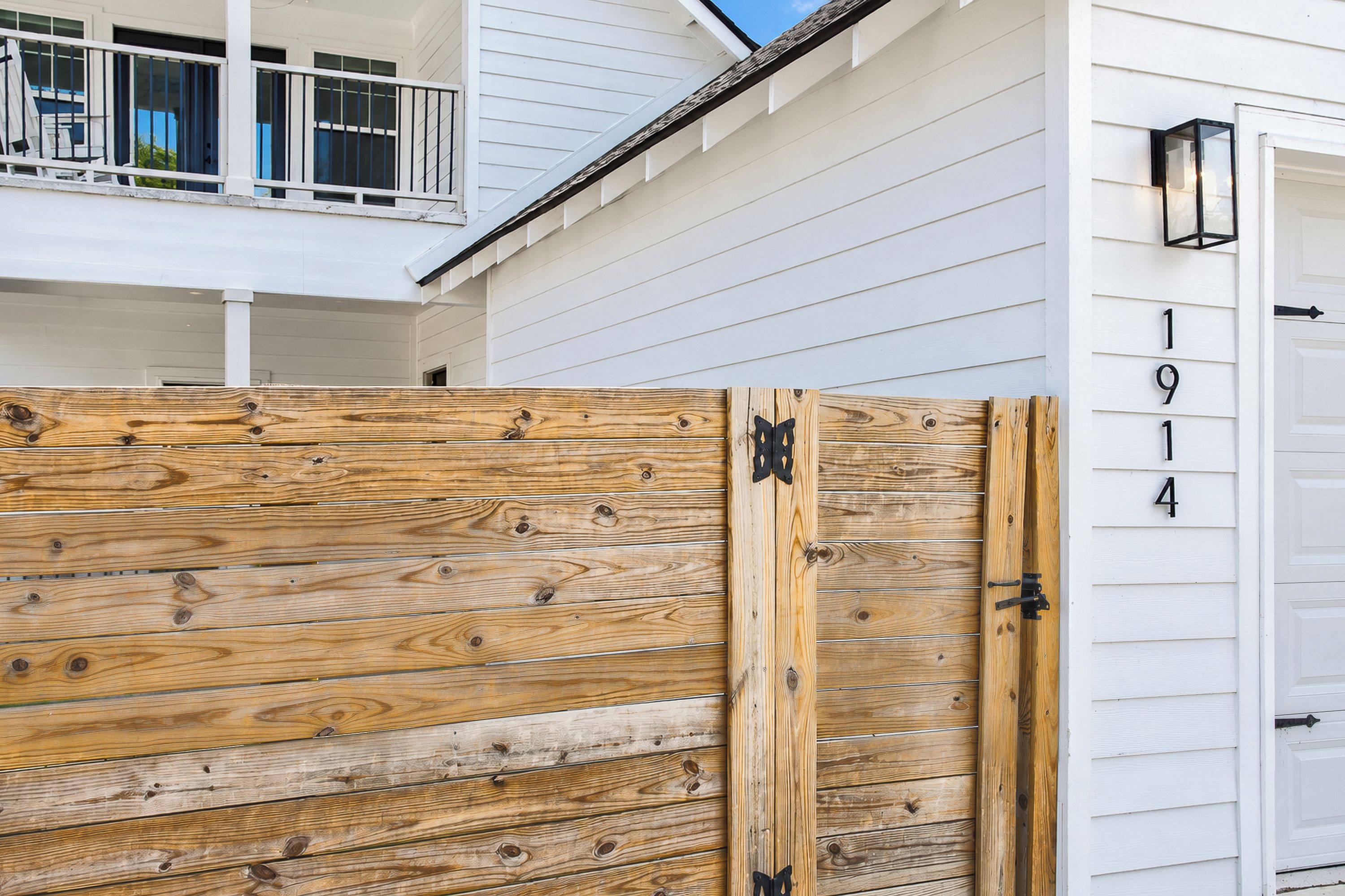 A wooden gate with a metal latch, a white house with a balcony, and a garage door with the numbers '1914' visible.