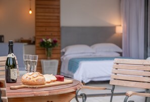 A dining table with a bottle of wine, glasses, a loaf of bread, and a small plate of cheese. A bed with white bedding and a wooden headboard is in the background. - Garden Room, Westmeston, Sussex (Westmeston)