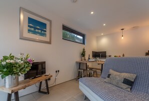 A modern living room with a sofa, a TV, a wooden table with a plant, and a kitchen area in the background. - Garden Room, Westmeston, Sussex (Westmeston)