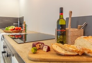 Una encimera de cocina con una botella de vino, una tabla de cortar con queso y fruta, y una cesta con pan y utensilios.