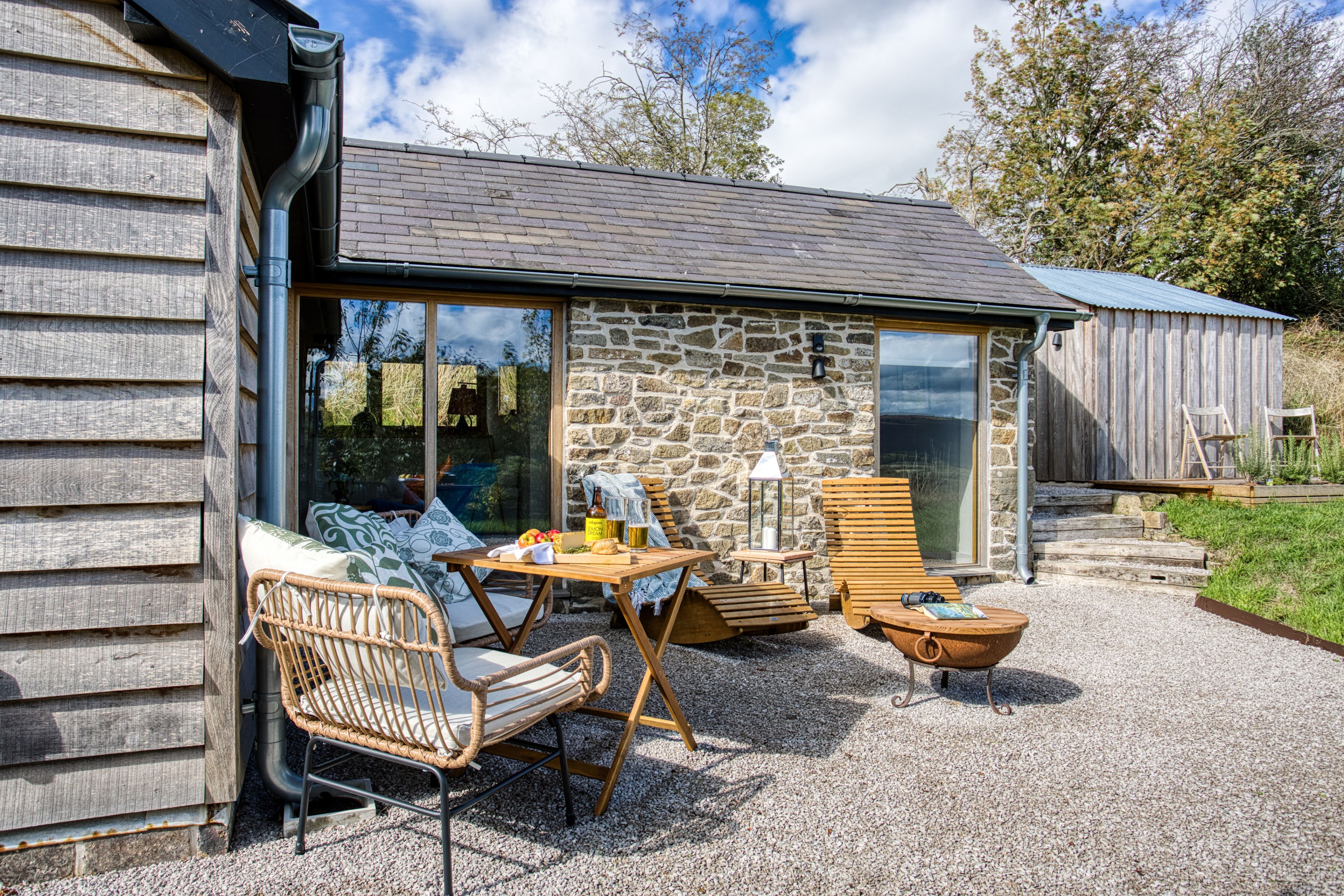A stone building with a large window, a wooden table, wicker chairs, and a gravel patio.