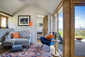 A cozy living room with a grey sofa, a blue armchair, and a patterned rug. There is a window with a view of the outdoors. - Little Barn, Minsterley, Shropshire (Minsterley)