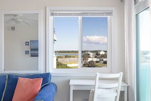A room with a view of a beach and houses, a ceiling fan, a small table, and a blue couch. - SeaSpray 305 | Oceanview Condo w/ Pool (Atlantic Beach)