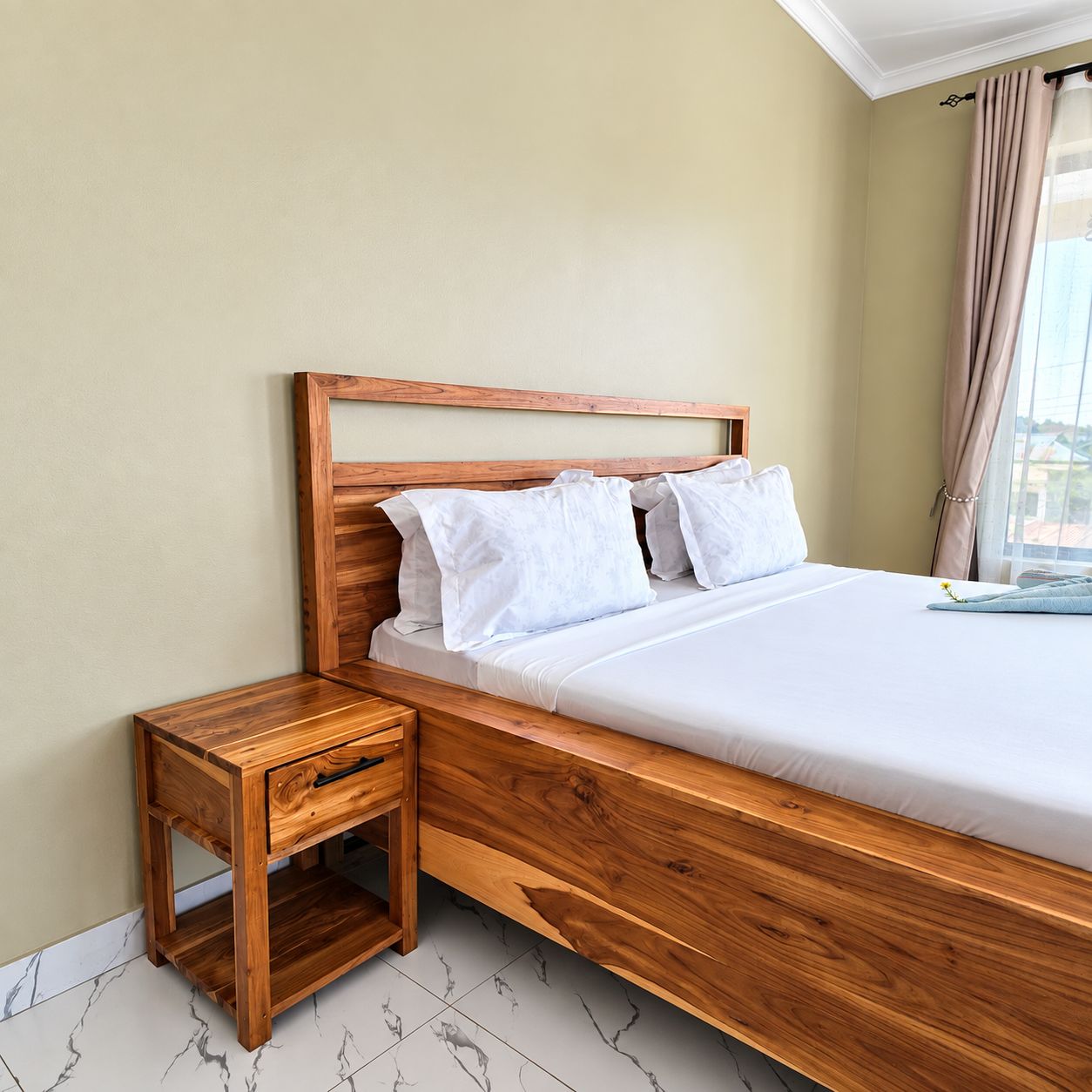 A neatly made bed with a wooden headboard and white bedding, a small wooden nightstand with a drawer, and a window with beige curtains.