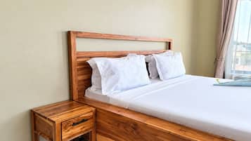 A neatly made bed with a wooden headboard and white bedding, a small wooden nightstand with a drawer, and a window with beige curtains.
