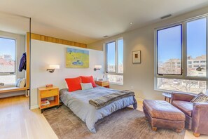 A bedroom with a bed, a nightstand, a reading chair, large windows, and artwork on the wall. - L'Avenir on Oak | Steps from Old Town Square (Fort Collins)