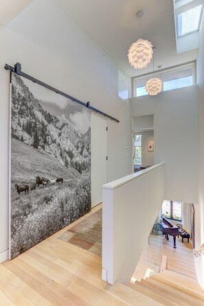 En modern inredning med ett stort väggmonterat konstverk som föreställer ett bergigt landskap, ett piano och en trappa som leder upp till en övre våning.
