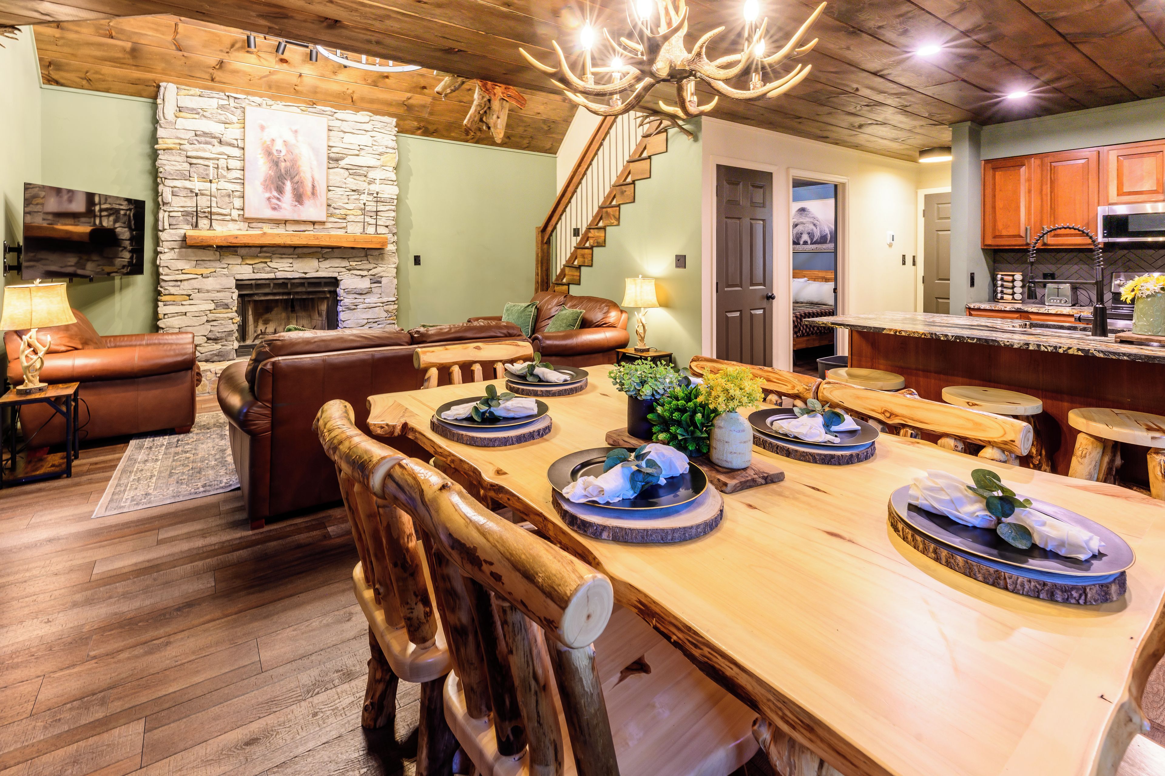 A rustic interior with a stone fireplace, leather furniture, a wooden dining table with plates, and a staircase.