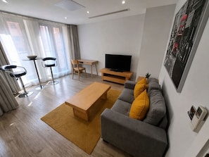 A modern living room with a sofa, coffee table, TV, and bar stools. - City Court - Stylish 1BR Home in the City (London)